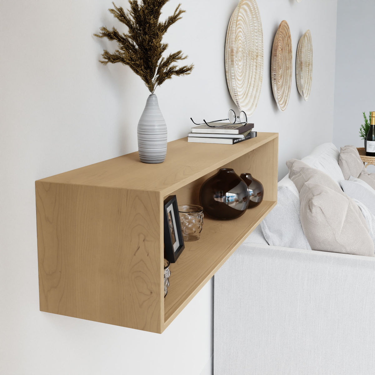 A Krovel Floating Display Case in Maple is mounted above a white sofa, showcasing a striped vase with dried flowers, two brown glass vases, books, a picture frame, and small decor. Woven wall art hangs on the wall behind the sofa.