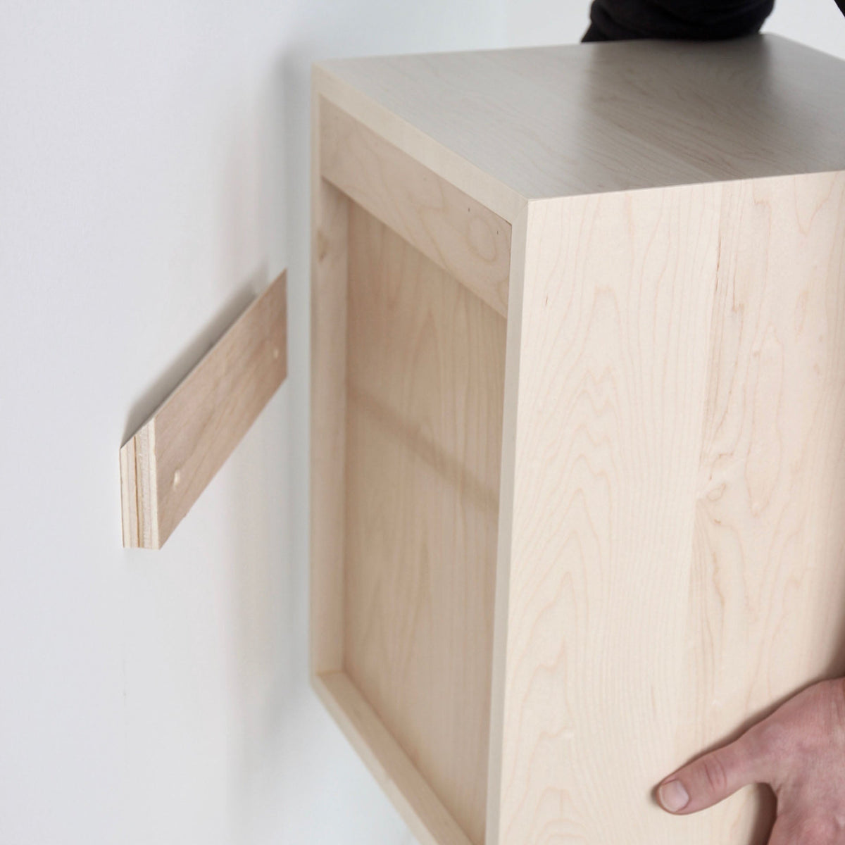 A person mounts the Krovel Floating Display Case in Maple to a wall with a French cleat—one piece on the wall, the other on the case—just like installing floating shelves.