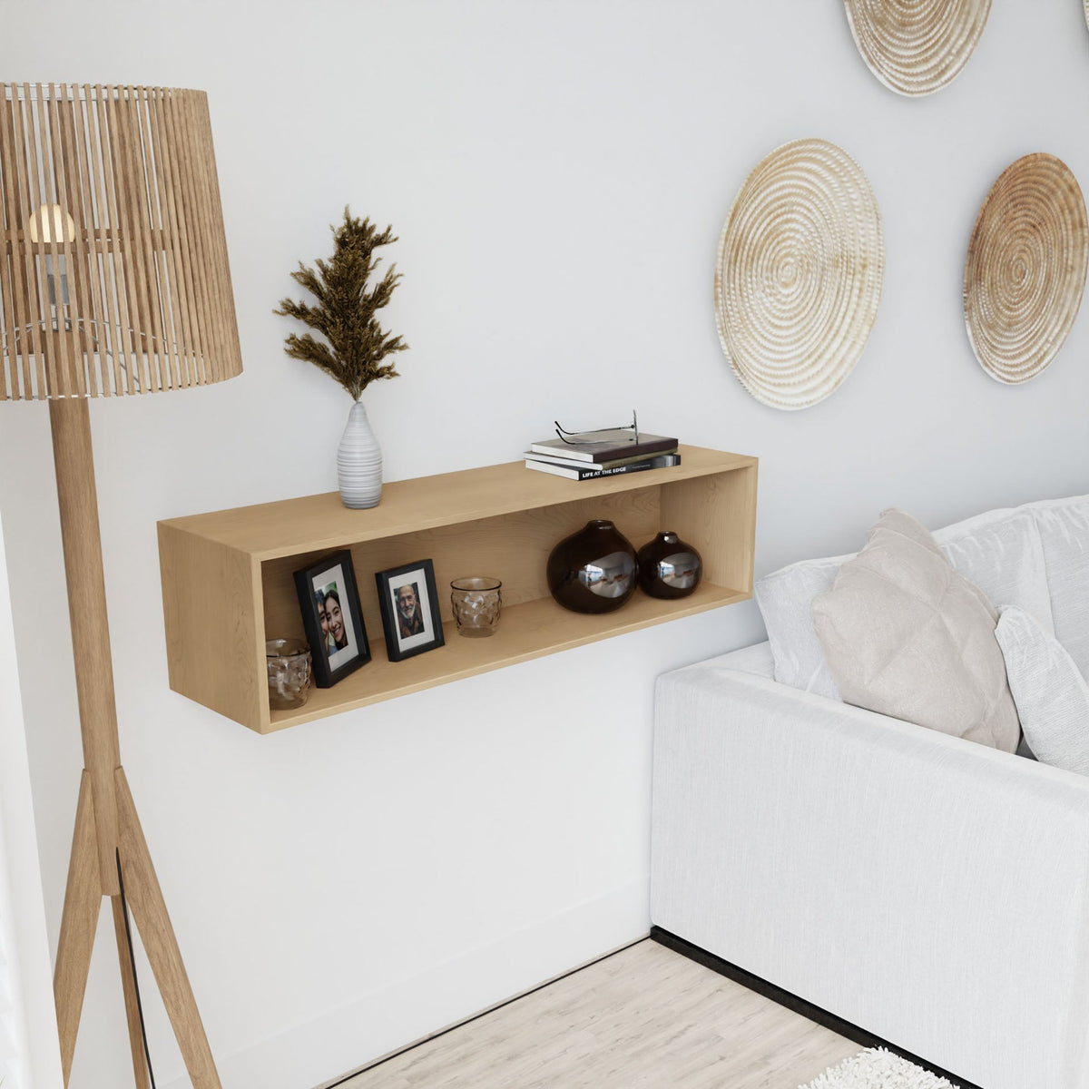 A modern living room corner features a handmade Hard Maple floor lamp, a Krovel Floating Display Case in Maple with a vase of dried flowers, framed photos, decorative bowls, and candles beside a white sofa and woven wall art.