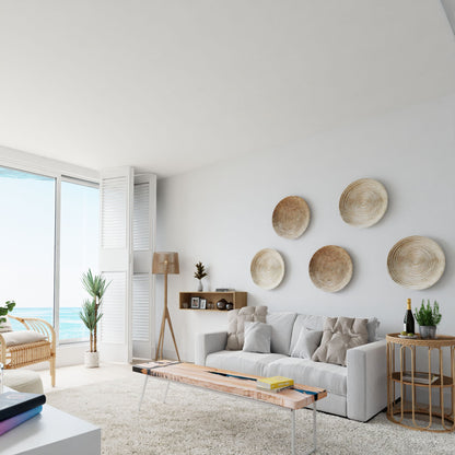 A bright, modern living room with a white sofa, solid hardwood coffee table, round wicker wall art, large ocean-view window, and a cozy coastal vibe. Decorative items are displayed on Krovel Hardwood Floating Display Cases.