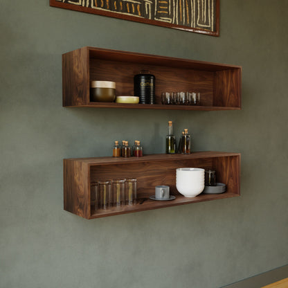 Two Krovel Hardwood Floating Display Cases are mounted on a green wall. The top case highlights elegant glassware and a coffee container, while the bottom displays glasses, a mug, and neatly stacked white plates.
