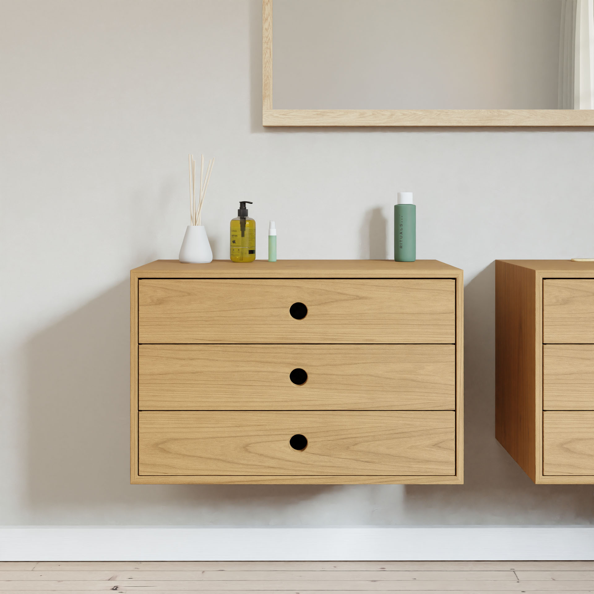 A Krovel Floating Dresser in Cherry, featuring three drawers, is mounted on a pale wall, resembling hand-finished hardwood furniture. Skincare bottles and a reed diffuser vase adorn the top. A partially visible wooden-framed mirror hangs above, while light wood flooring completes the scene.