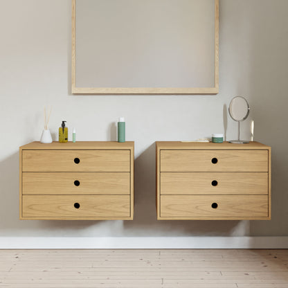 Two Krovel Floating Dressers in Cherry, each with three drawers, are centered under a large mirror against a pale wall. Bottles, a reed diffuser, and a circular mirror adorn the dressers. This hand-finished hardwood furniture complements the light wood flooring elegantly.