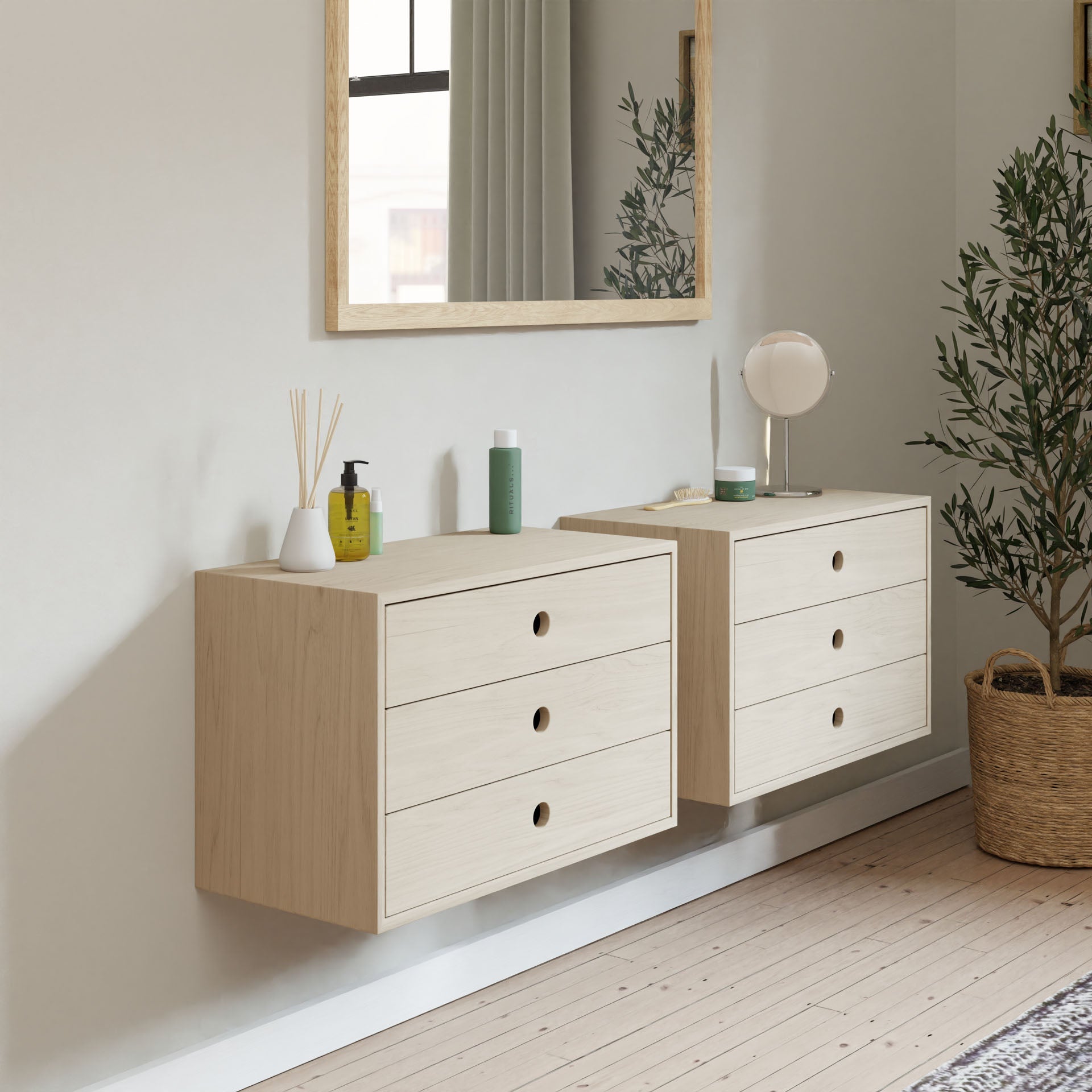 A minimalist room showcases two Krovel Floating Dressers in Maple, each featuring three drawers with circular handles and soft-close slides. A large mirror hangs above, while a potted plant sits in the corner, and small items like bottles and a lamp adorn the dressers.