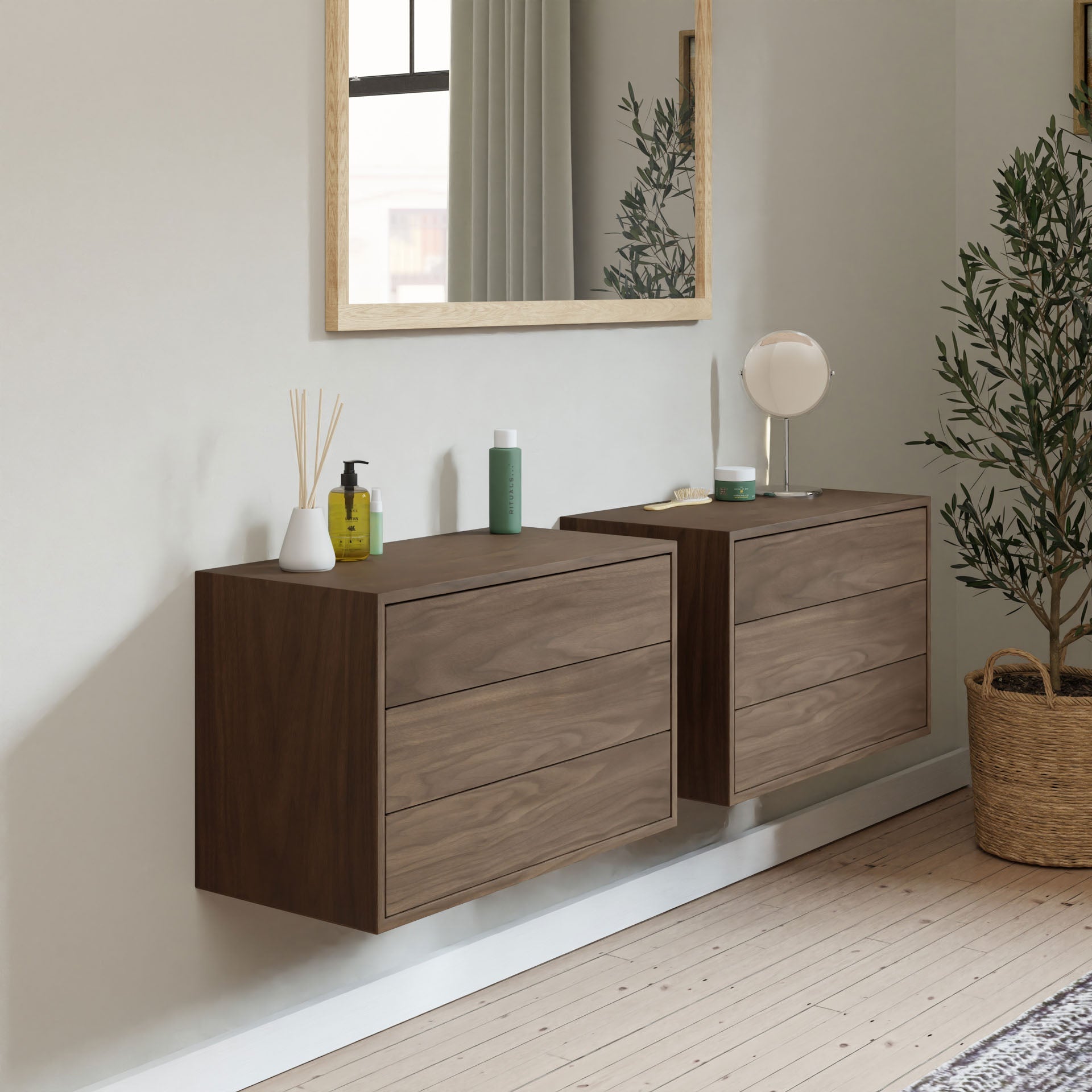 A minimalist room features two Krovel Floating Dressers in Walnut, with a large mirror above and various toiletries on top. A potted plant adorns the wooden floor beside the cabinets, showcasing a modern and tidy style.