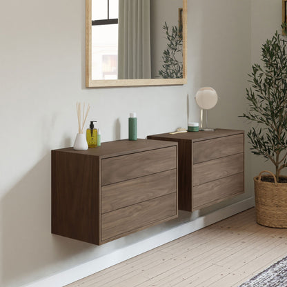 A minimalist room features two Krovel Floating Dressers in Walnut, with a large mirror above and various toiletries on top. A potted plant adorns the wooden floor beside the cabinets, showcasing a modern and tidy style.