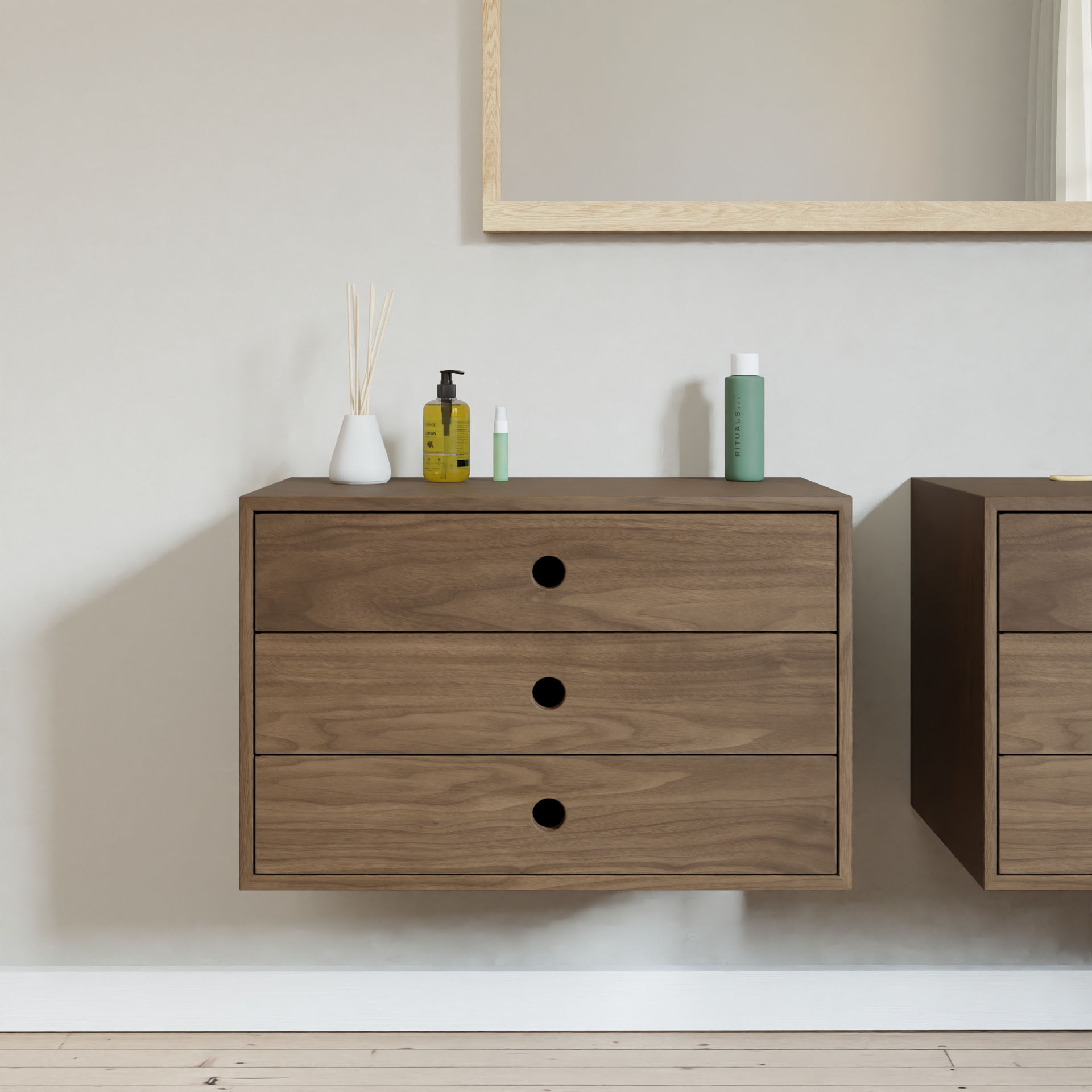 The Krovel Floating Dresser in Walnut is wall-mounted and features three drawers with black circular knobs. It displays a white vase with reed sticks and various bottles on top, with a partially visible wooden-framed mirror above. Enjoy its luxury soft-close premium slides for effortless use.