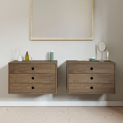 Two Krovel Floating Dressers in walnut, each with three drawers and premium soft-close slides, are mounted against a light wall. A large mirror hangs above, while various toiletries and a small round mirror rest on the dressers. Light wood flooring is visible below.