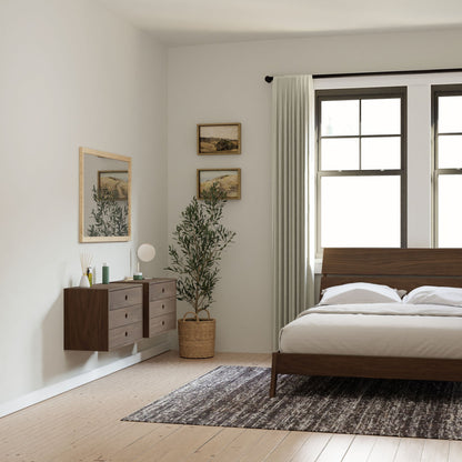 A minimalist bedroom features a wooden bed, white bedding, and a textured rug. Krovel's Floating Dresser in Walnut with soft-close slides sits below a mirror and two framed artworks. A potted plant adorns the window with light curtains, complementing light-colored walls and flooring.