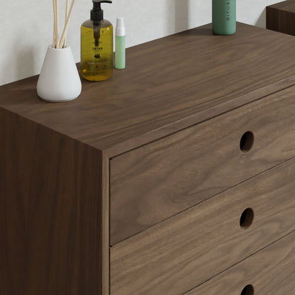A Krovel Floating Dresser in Walnut is placed against a wall. Topped with a white vase with reed diffusers, a yellow pump bottle, and a green spray bottle, it features soft-close premium slides and circular cut-out handles.