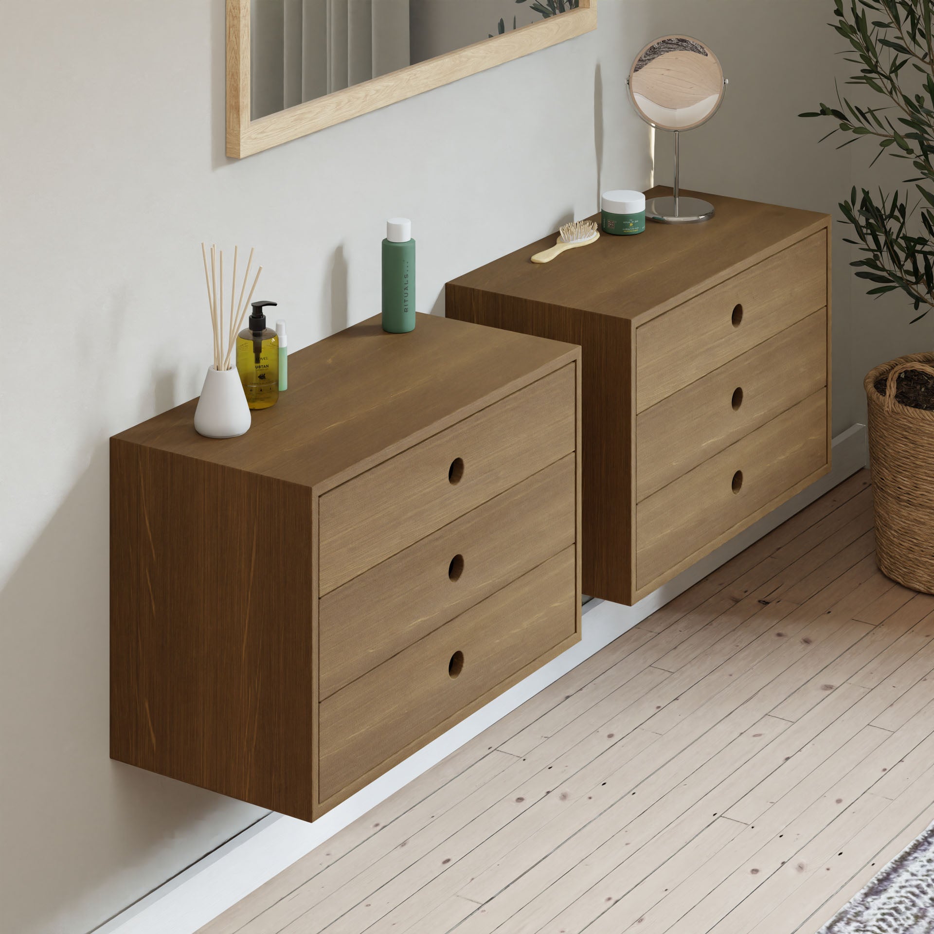 Two Krovel Floating Dressers in White Oak are mounted on a light wall, each holding grooming items like a green bottle, round mirror, and reed diffuser. A potted plant sits nearby on the wooden floor with another mirror hanging above.