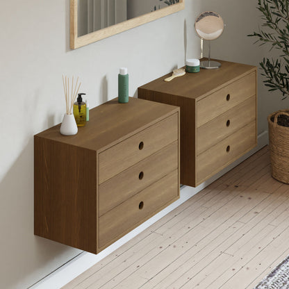 Two Krovel Floating Dressers in White Oak are mounted on a light wall, each holding grooming items like a green bottle, round mirror, and reed diffuser. A potted plant sits nearby on the wooden floor with another mirror hanging above.