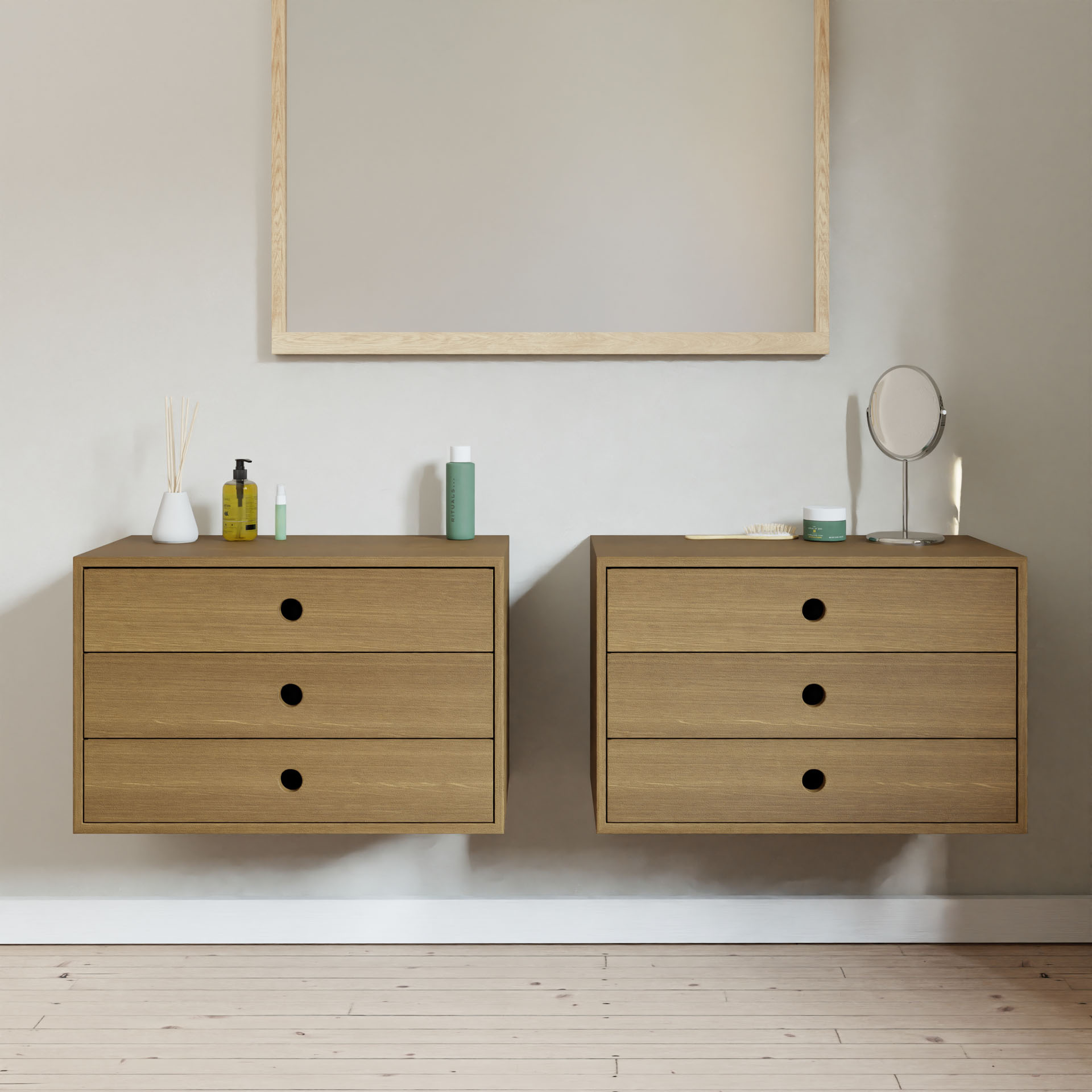 A minimalist bedroom features two Krovel Floating Dressers in White Oak, each with three drawers. The top holds personal care items like bottles and a small mirror. A large blank canvas hangs above on the light-colored wall, while light wooden flooring enhances the serene atmosphere.