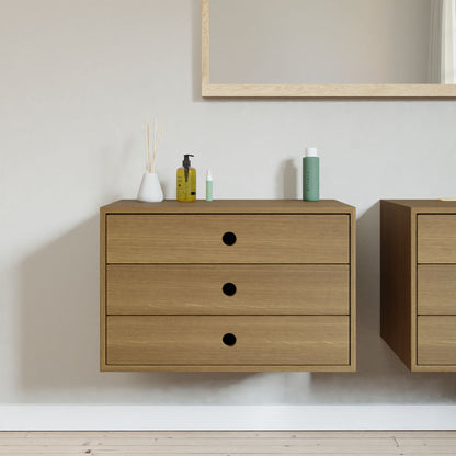 In this minimalist room, two Krovel Floating Dressers in White Oak adorn a pale wall. A mirror is mounted above one dresser, which holds a vase with sticks and toiletries. The light wood floor enhances the serene atmosphere.