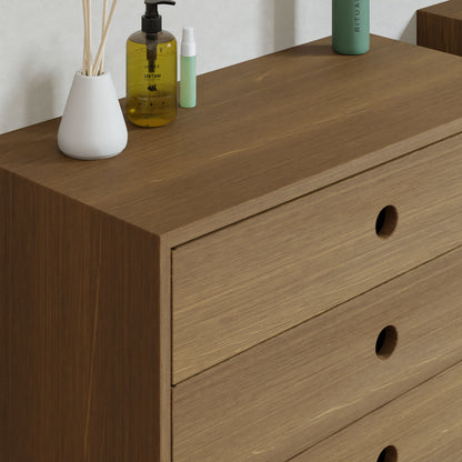 A Krovel Floating Dresser in white oak with three drawers and simple round handles holds a reed diffuser, yellow bottle with pump, and green cylindrical container, all set against a plain white wall.