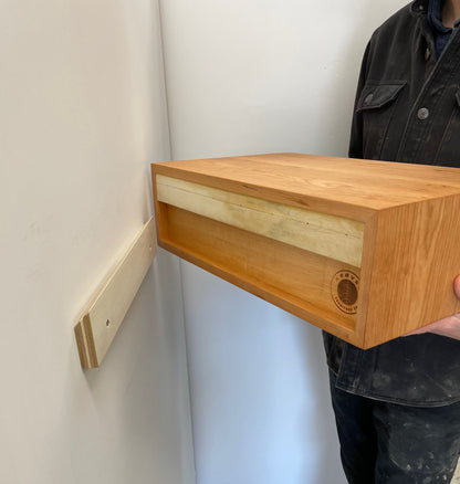 A person holds a Krovel Hardwood Floating Nightstand Shelf near a light-colored wall. A wooden mounting bracket is attached to the wall, ready for installation of the shelf unit.