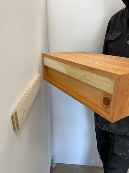 A person holds the Krovel Hardwood Floating Single Nightstand, featuring a wall-mount bracket for easy installation. This solid hardwood shelf has a smooth finish and displays the maker’s logo on the lower right corner.