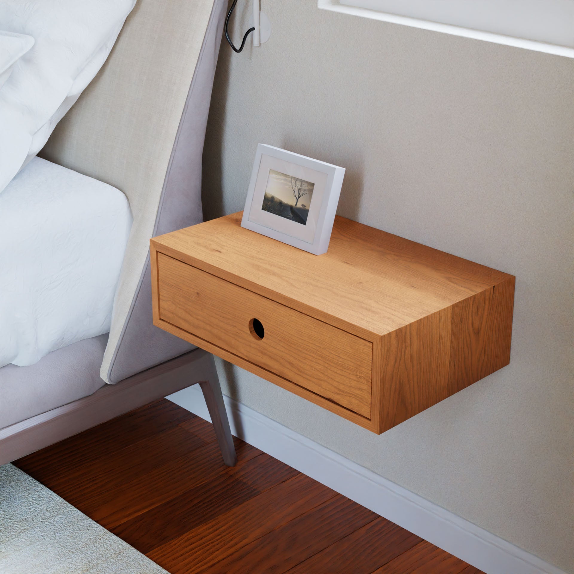 The Krovel Hardwood Floating Single Nightstand, a handmade wall-mounted piece with one drawer, sits beside a bed and holds a small framed photo. Solid hardwood construction matches the wood floor; part of a window is in view.