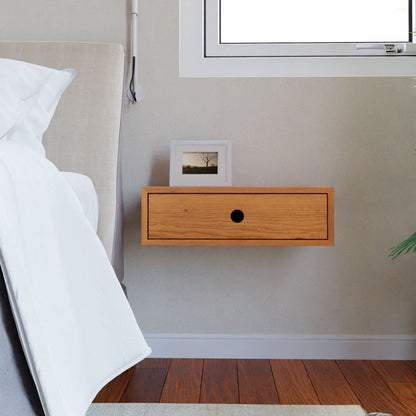 A modern bedroom features the Krovel Hardwood Floating Single Nightstand, holding a small framed photo next to a white bed and window above, with a plant partially visible on the right.