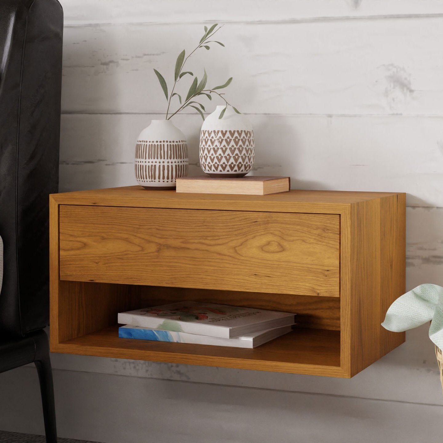 The Krovel Hardwood Double Floating Nightstand, crafted from solid hardwood with a drawer and open shelf, is mounted on a white wood-paneled wall by a black chair and holds two vases, a tray, and books.