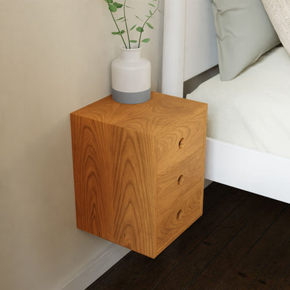 The Krovel Hardwood Triple Floating Nightstand, crafted from solid hardwood, is wall-mounted beside the bed and topped with a white vase of green leafy stems, perfectly complementing the dark wood floor.