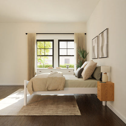 A minimalist bedroom showcases a white bed with neutral bedding alongside a Krovel Floating Nightstand in Cherry, crafted from solid wood. A large window with beige curtains fills the room with natural light, while modern artwork and a potted plant enhance the decor. A soft rug is spread out on the dark wooden floor.