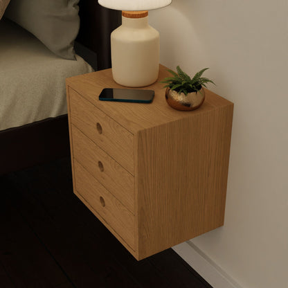 The Krovel Hardwood Triple Floating Nightstand, made from solid hardwood, features three drawers and holds a lamp, gold potted plant, and smartphone next to a bed with beige bedding.