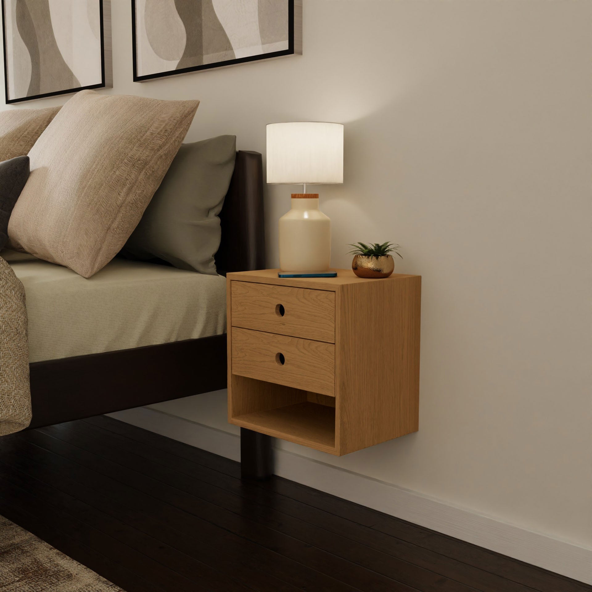 A modern bedroom showcases the Krovel Floating Nightstand in Cherry, secured with a French cleat and crafted from solid wood. Two drawers offer storage space, while the top is adorned with a lamp, a small potted plant, and a book. The bed features beige pillows resting on a brown frame, accentuated by abstract art hanging on the wall above.#configuration_double-drawer-single-shelf