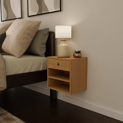 A modern bedroom displays a Krovel Floating Nightstand in cherry, elegantly mounted with a French cleat. It includes a drawer and two open shelves, adorned with a beige lamp and a small potted plant. The corner of the bed, dressed in neutral linens, rests on the dark wooden floor beneath abstract artwork above.