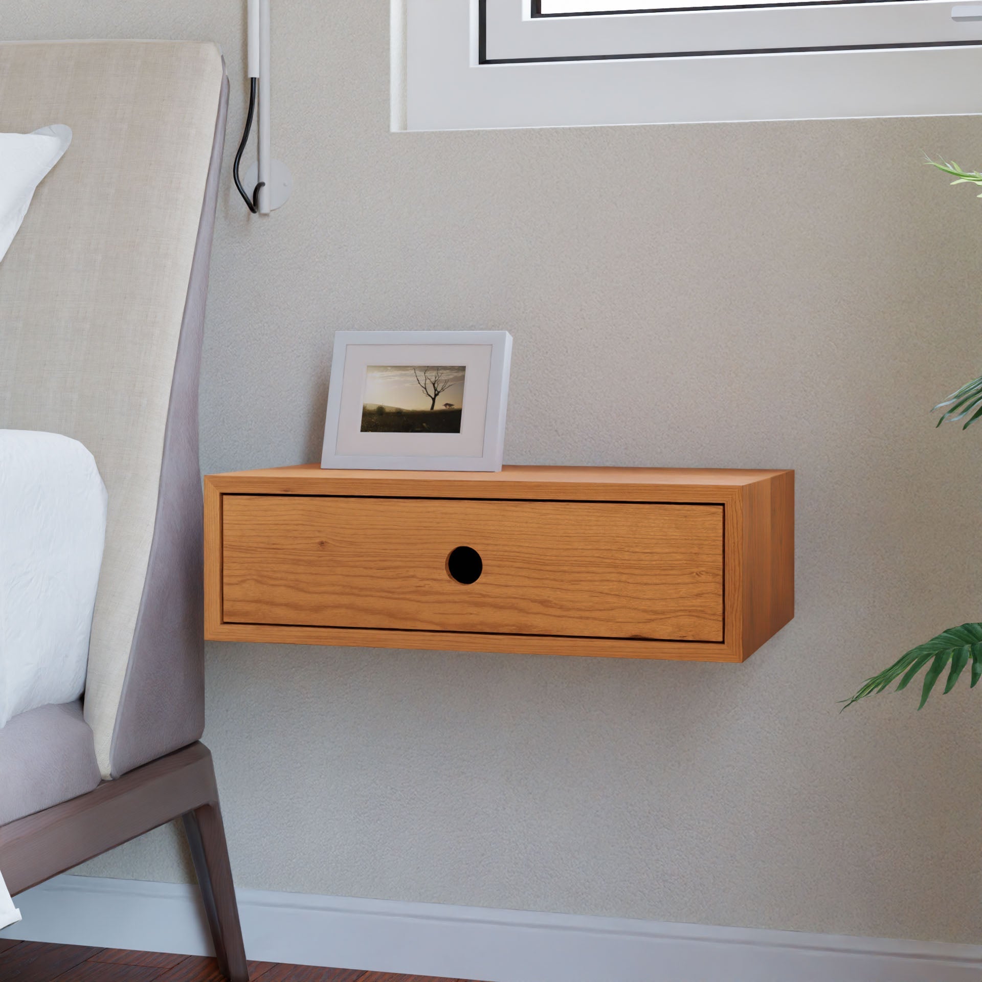 In a minimalist bedroom scene, a Krovel Floating Nightstand in Cherry is featured. It boasts elegant craftsmanship with a discreet French cleat for seamless installation. The piece includes a single drawer, and atop it rests a framed photo of a tree silhouette, complemented by glimpses of a bed and nearby plant.#configuration_single-drawer