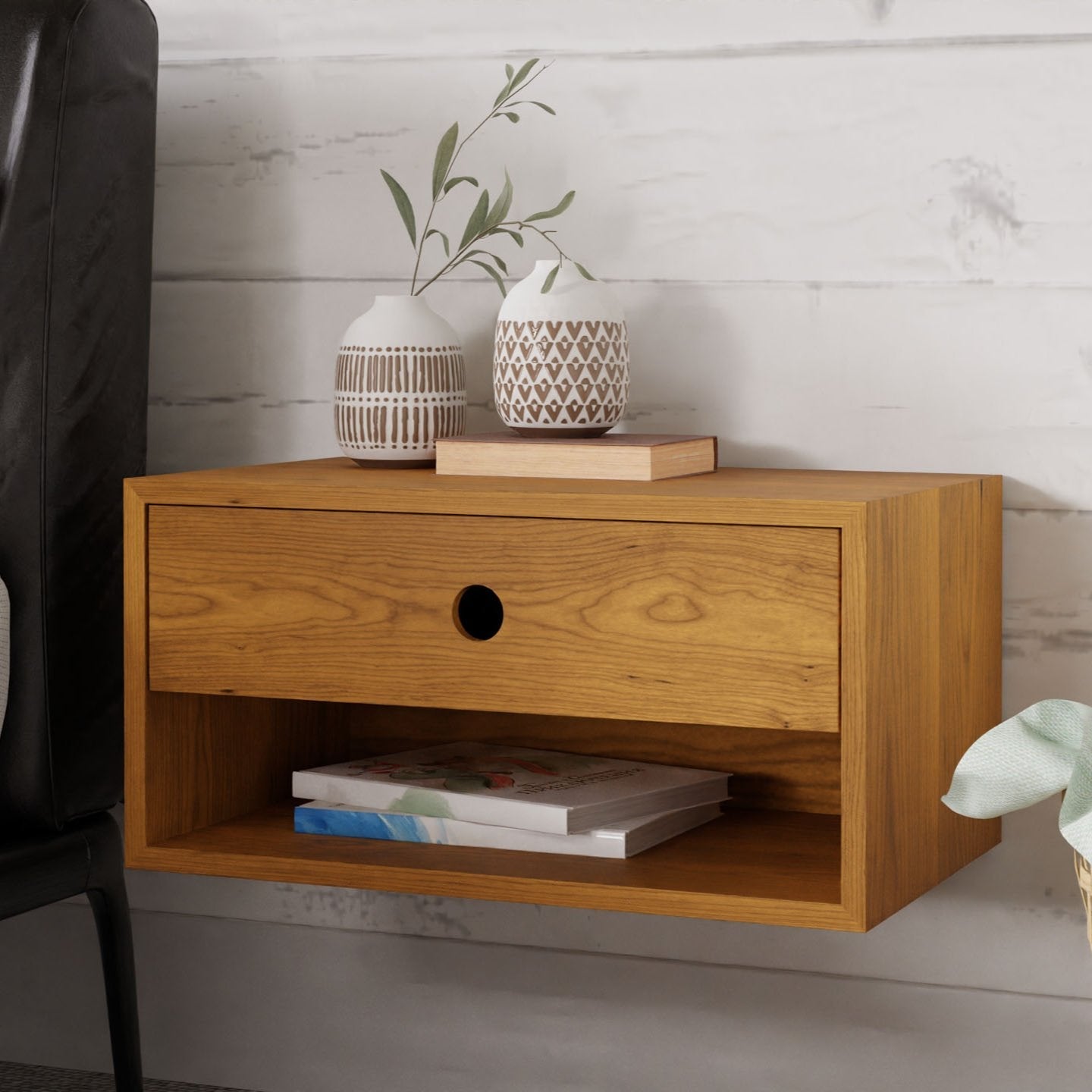 A sophisticated Floating Nightstand in Cherry, crafted by Krovel with a single drawer and open shelf, is elegantly mounted using a French cleat. A patterned vase containing green branches and a small decorative object adorn the top, while a book rests on the lower shelf. Nearby, presence of a black chair along with part of a plant completes this tranquil setting.#configuration_single-drawer-single-shelf