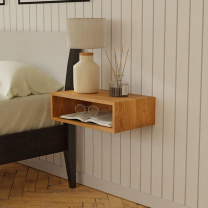 A floating nightstand in Cherry from Krovel, crafted to perfection, features an open shelf holding a closed book and glasses. A ceramic lamp and diffuser grace the top, while the backdrop of a grooved white wall enhances its elegance. Part of a bed with pristine white linens is visible on the left, all safely secured with a French cleat system.