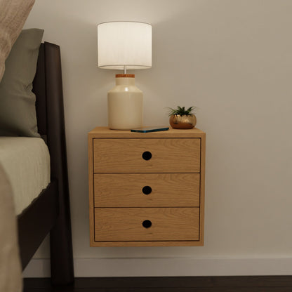 A Floating Nightstand in Cherry by Krovel is mounted on the wall beside a bed, secured with a discreet French cleat. The minimalist design is complemented by a ceramic-based lamp with a white shade, accompanied by a small plant and smartphone atop its three sleek drawers.