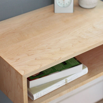 A close-up of the Krovel Hardwood Floating Nightstand Shelf showcases its open shelf with two books, while the surface holds a small round white container and a square clock, all set against a blue-gray wall.