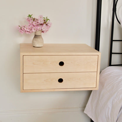 The Krovel Hardwood Double Floating Nightstand, featuring two drawers with round cut-out handles, is mounted on a white wall. A small vase with pink flowers rests on top, beside a bed dressed in crisp white bedding.