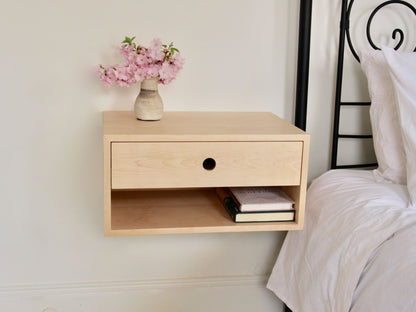 The Krovel Hardwood Double Floating Nightstand, handmade from solid hardwood, has a drawer and open shelf with two books and a small vase of pink flowers, mounted on a white wall beside a bed with crisp white bedding.