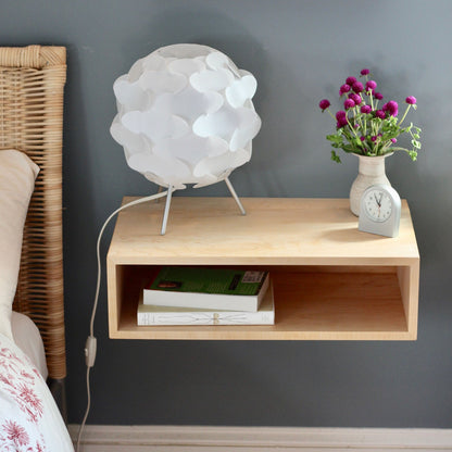 The Krovel Hardwood Floating Nightstand Shelf displays a modern white lamp, a small vase of purple flowers, a gray clock, and two books. A gray wall and part of a wicker bedframe are visible on the left.