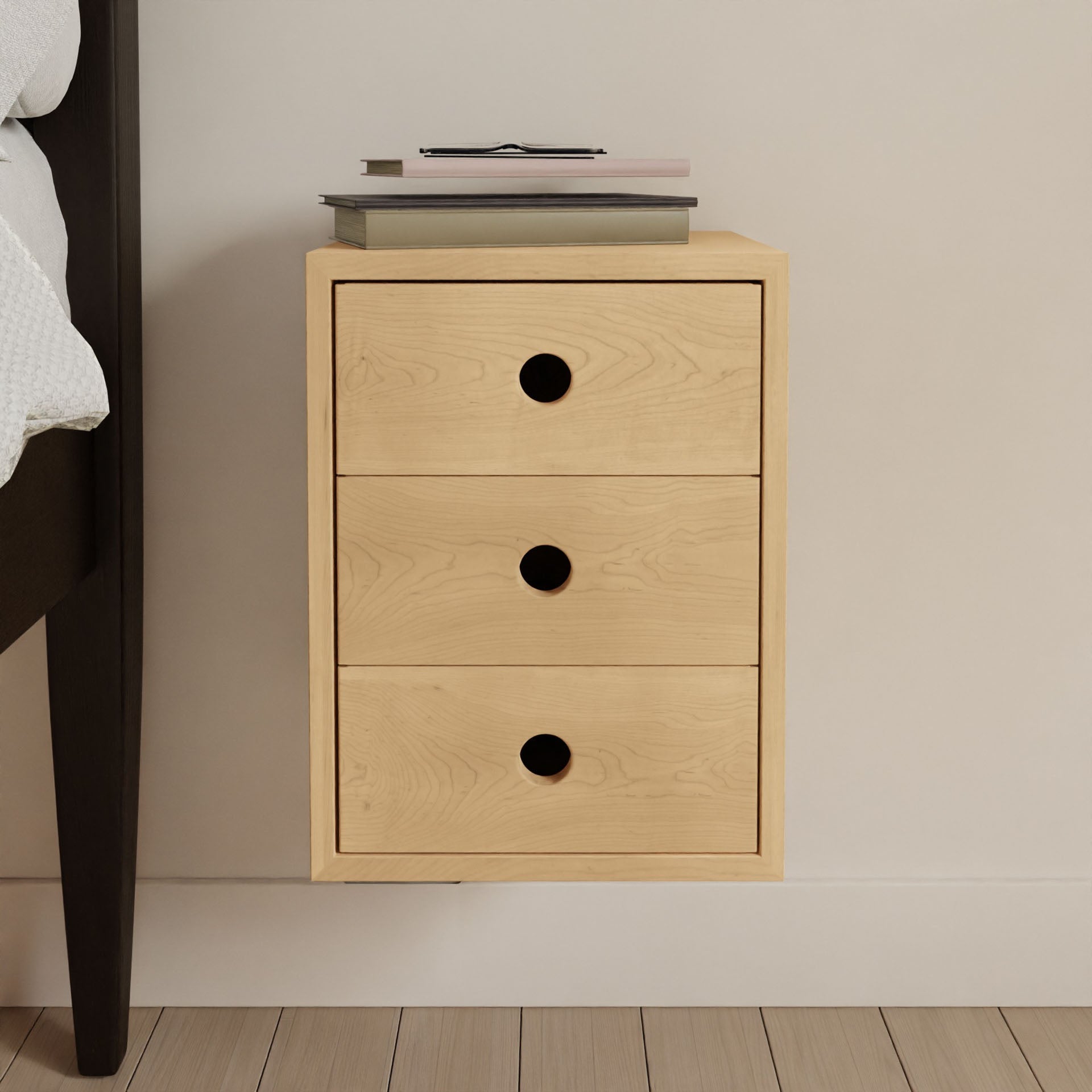 The Krovel Hardwood Triple Floating Nightstand is a handmade, wall-mounted unit in light solid hardwood with three drawers and round black handles. Shown beside a bed on matching wood floors, it holds books and a closed laptop on top.