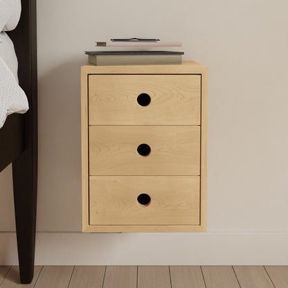 The Krovel Hardwood Triple Floating Nightstand is a handmade, wall-mounted unit in light solid hardwood with three drawers and round black handles. Shown beside a bed on matching wood floors, it holds books and a closed laptop on top.