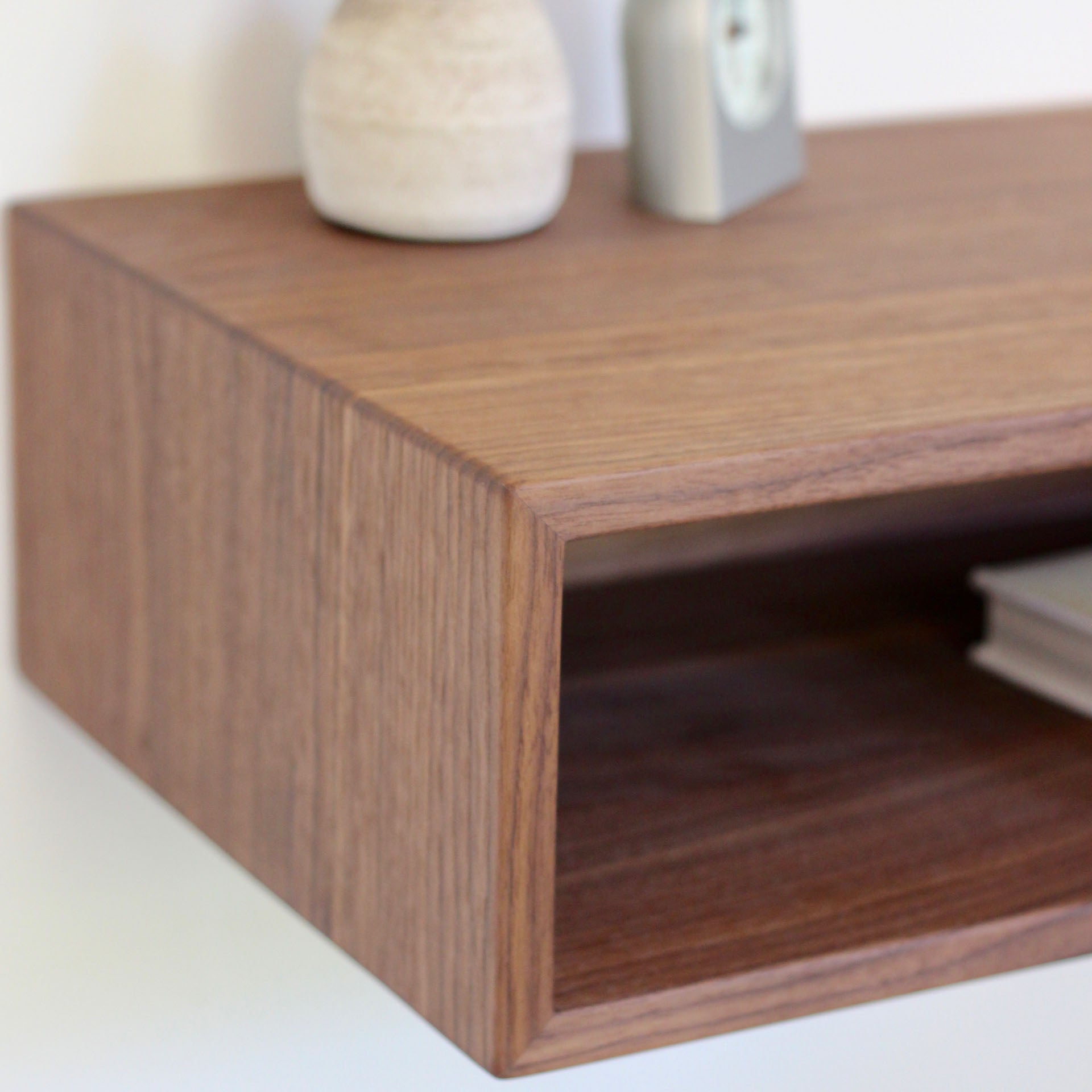 Close-up of the Krovel Hardwood Floating Nightstand Shelf, mounted on a white wall, featuring a small beige vase, square gray clock, and part of a book displayed on and inside the shelf.