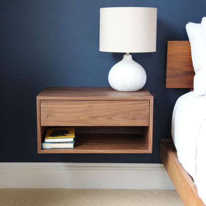 The Krovel Hardwood Double Floating Nightstand, handcrafted from solid hardwood, features a drawer and open shelf with two books. A white ceramic lamp with a beige shade rests on top beside a wooden bed against a dark blue wall.