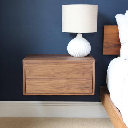 The Krovel Hardwood Double Floating Nightstand, handmade from solid hardwood with two drawers, is mounted on a dark blue wall beside a bed and topped with a textured white ceramic table lamp.