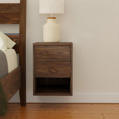 The Krovel Hardwood Triple Floating Nightstand, crafted from solid hardwood, features two drawers and an open shelf. It holds a cream-colored lamp beside a wooden bed frame on a wood floor.