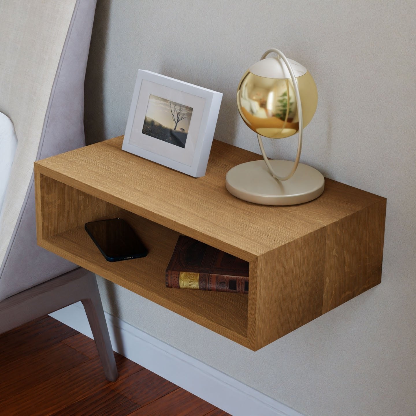 Perched against a light-colored wall beside a chair on hardwood flooring, the minimalist décor features a Krovel Floating Nightstand in White Oak. This sleek piece is adorned with a small lamp, framed photograph, and two books on its surface, while a smartphone rests conveniently on the shelf below. 