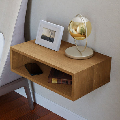 Perched against a light-colored wall beside a chair on hardwood flooring, the minimalist décor features a Krovel Floating Nightstand in White Oak. This sleek piece is adorned with a small lamp, framed photograph, and two books on its surface, while a smartphone rests conveniently on the shelf below. 