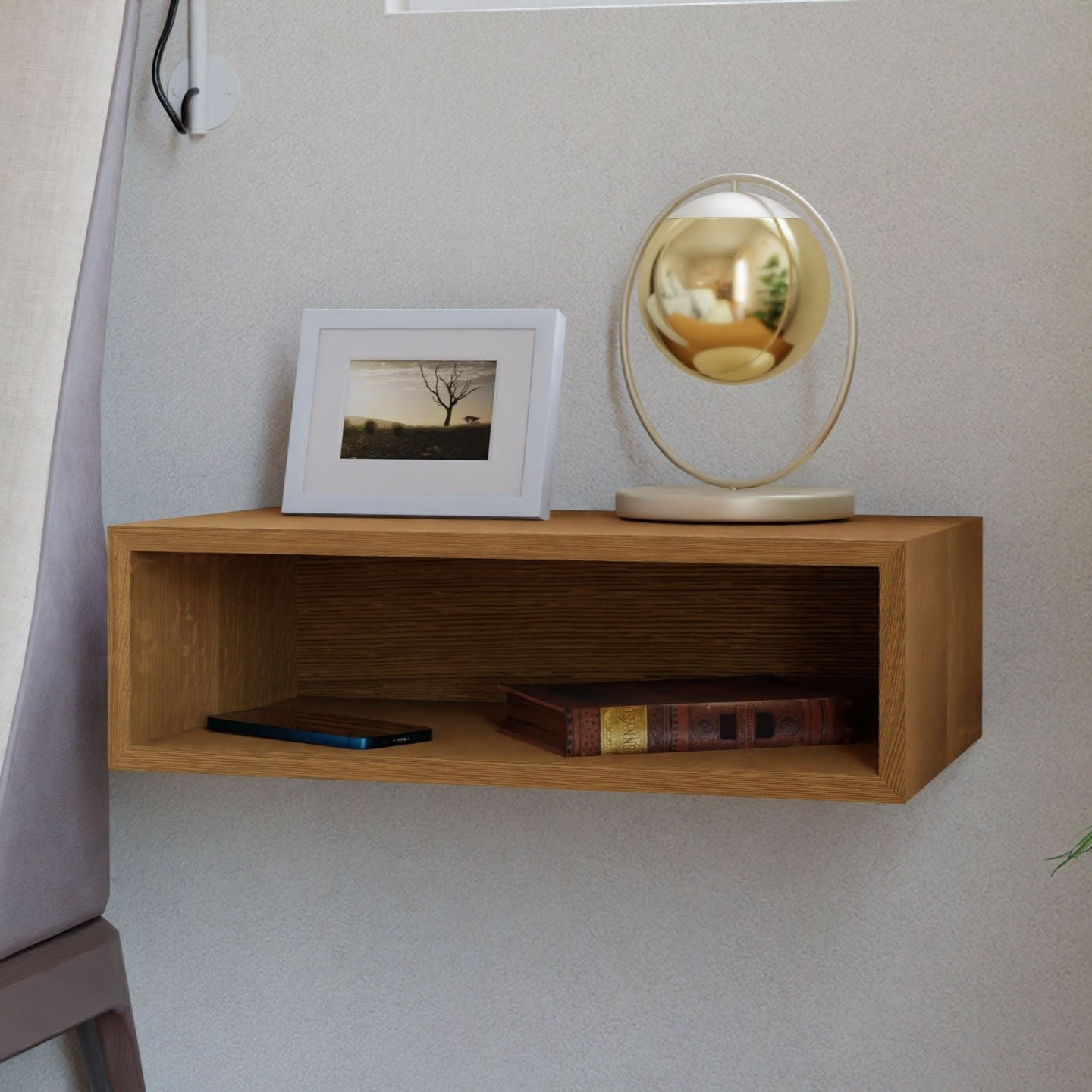 A Floating Nightstand in White Oak by Krovel is mounted on a light-colored wall, serving also as a floating shelf. It supports a framed photo of a tree and a gold circular mirror. Below, you can see a closed book and smartphone. An upholstered chair is partially visible on the left. 