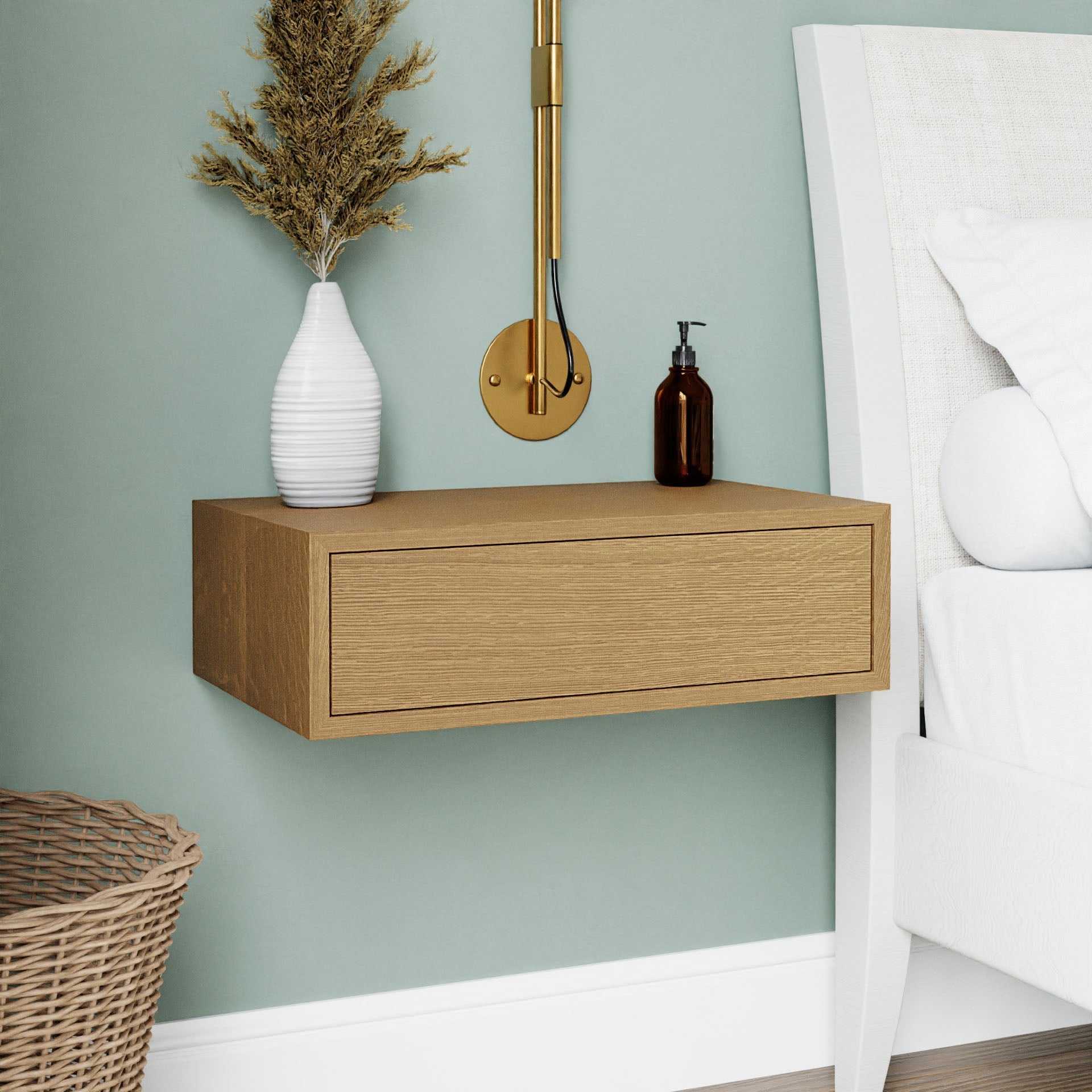 A modern bedroom scene highlights the Krovel Floating Nightstand in White Oak, adorned with a white vase containing dried flowers and a brown bottle. Above the nightstand, a gold wall-mounted lamp casts a warm glow, while a woven basket is placed on the green-painted floor. The nightstand features a soft-close drawer for quiet elegance. 