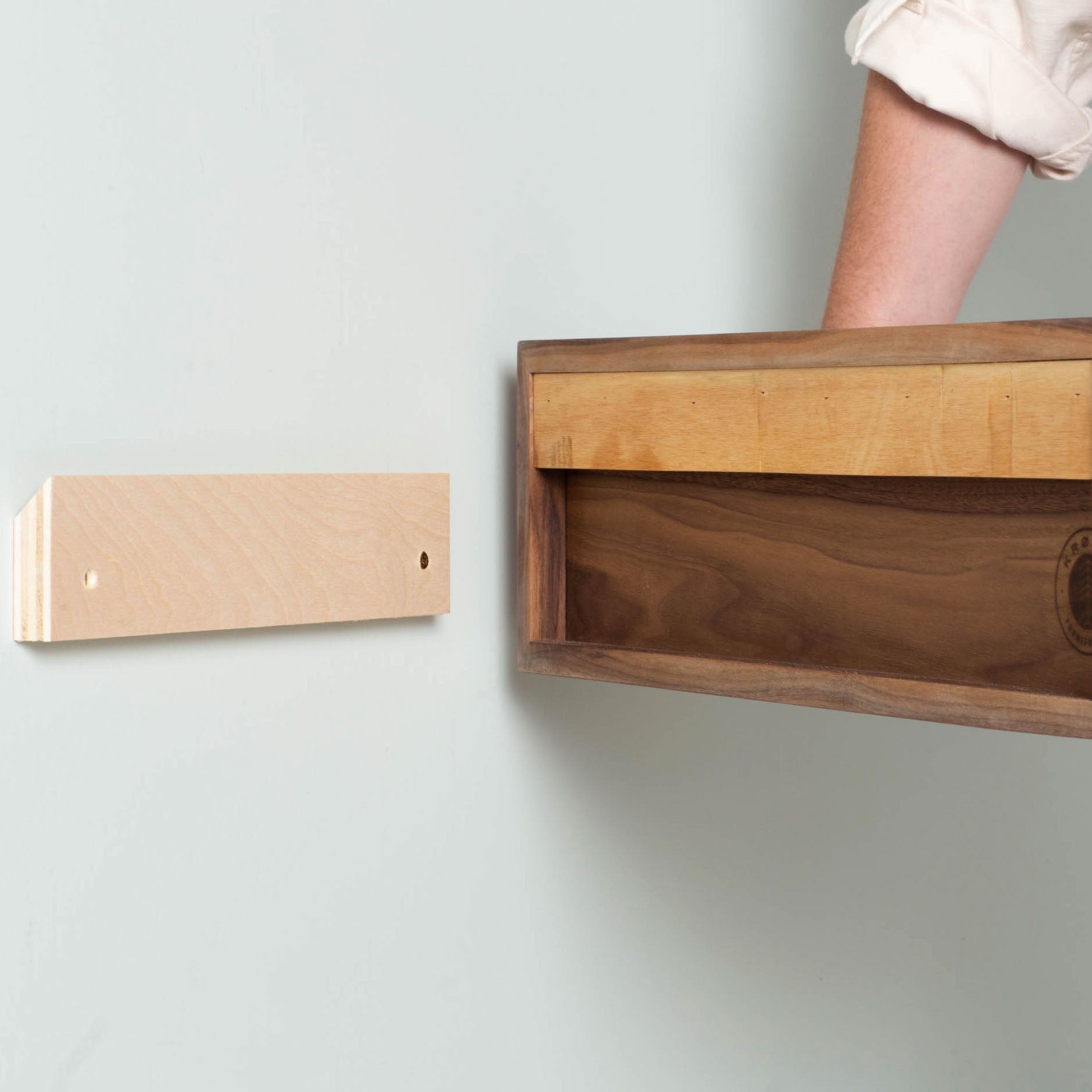 A person's arm is inserting a solid American White Oak bracket into the rectangular wooden shelf of Krovel's Floating Nightstand in White Oak. The wall-mounted bracket, featuring two screw holes, is attached to a light wall to ensure the shelf fits securely and seamlessly. 