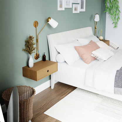 A modern bedroom showcases a white bed adorned with light blue and pink pillows, complemented by Krovel's Floating Nightstand in White Oak featuring brass lamps. A woven basket adds texture, and a green accent wall displays framed pictures. A plant brings in greenery, while a neutral rug enhances the wooden floor. 