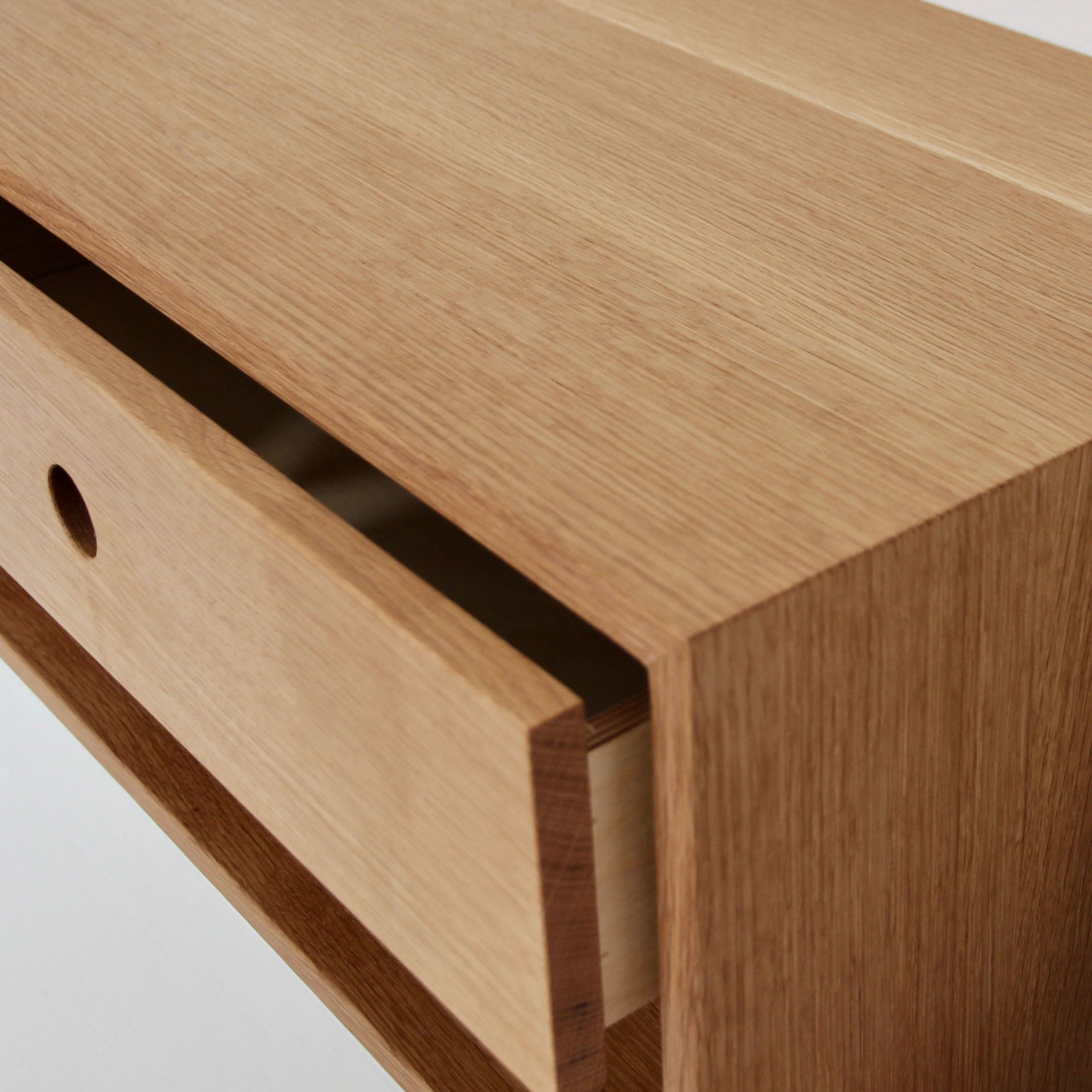 Close-up of the Krovel Floating Nightstand in White Oak, crafted from solid American White Oak, with a slightly open drawer revealing its inner compartment. The surface features a smooth, natural finish that highlights the wood grain. This soft-close drawer includes a circular cutout handle for easy access, making it an ideal addition to any room. 